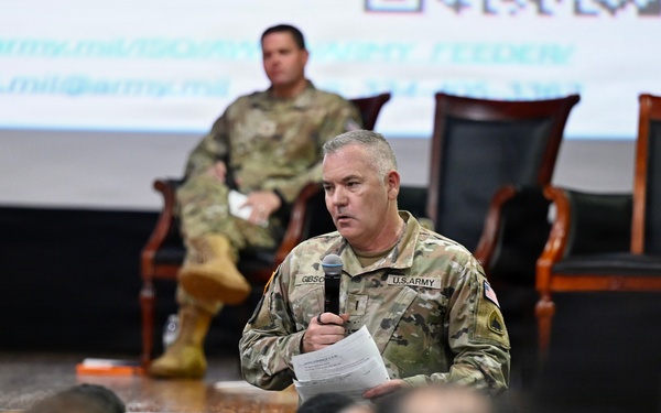 A U.S. Army soldier in OCP (Operational Camouflage Pattern) uniform is standing at the front of an auditorium-style room, speaking into a microphone while holding papers. A second soldier is seated on a chair on a raised stage in the background. The event appears to be a military briefing or town hall, with an audience of uniformed personnel visible in the foreground.