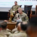 A U.S. Army soldier in OCP (Operational Camouflage Pattern) uniform is standing at the front of an auditorium-style room, speaking into a microphone while holding papers. A second soldier is seated on a chair on a raised stage in the background. The event appears to be a military briefing or town hall, with an audience of uniformed personnel visible in the foreground.
