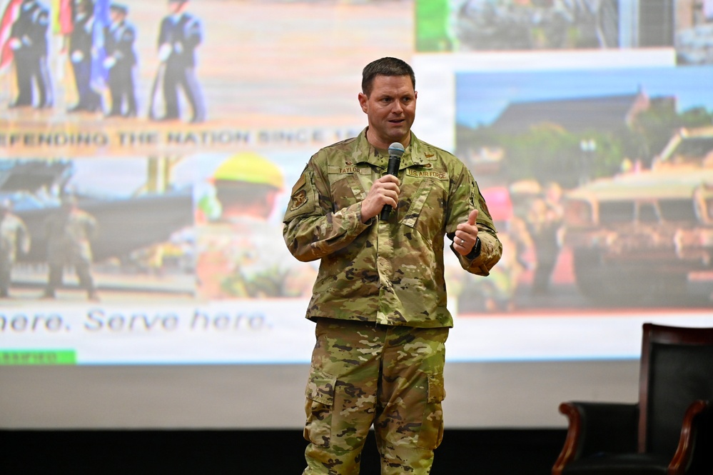 A U.S. Air Force servicemember named Taylor is speaking at a presentation or briefing, holding a microphone on a stage. He is wearing OCP (Operational Camouflage Pattern) uniform with a 'U.S. AIR FORCE' tape and name tape 'TAYLOR' visible. Behind him is a large projection screen displaying a collage of military images with text reading 'DEFENDING THE NATION SINCE...' and 'here. Serve here...'