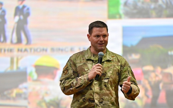 A U.S. Air Force servicemember named Taylor is speaking at a presentation or briefing, holding a microphone on a stage. He is wearing OCP (Operational Camouflage Pattern) uniform with a 'U.S. AIR FORCE' tape and name tape 'TAYLOR' visible. Behind him is a large projection screen displaying a collage of military images with text reading 'DEFENDING THE NATION SINCE...' and 'here. Serve here...'
