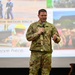 A U.S. Air Force servicemember named Taylor is speaking at a presentation or briefing, holding a microphone on a stage. He is wearing OCP (Operational Camouflage Pattern) uniform with a 'U.S. AIR FORCE' tape and name tape 'TAYLOR' visible. Behind him is a large projection screen displaying a collage of military images with text reading 'DEFENDING THE NATION SINCE...' and 'here. Serve here...'