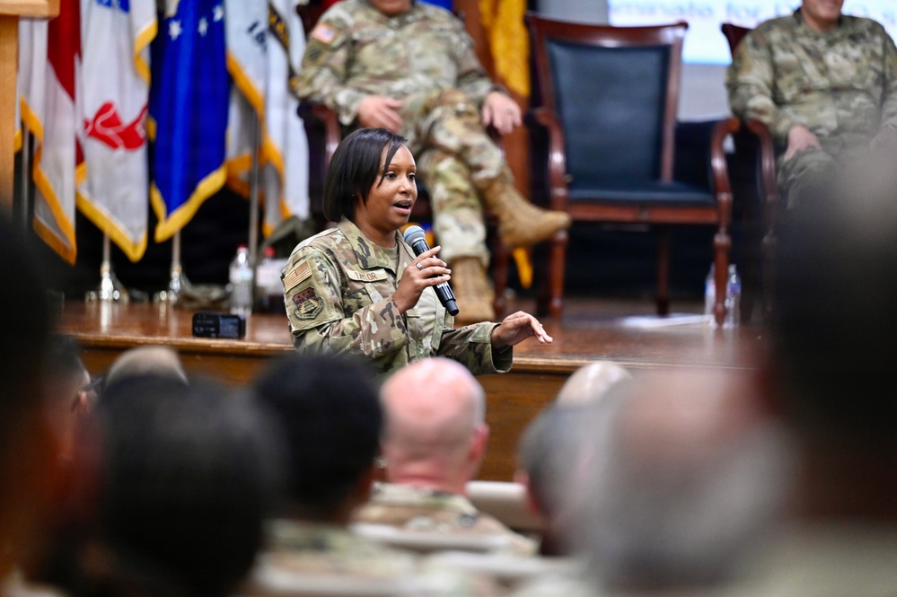 A female military service member in OCP (Operational Camouflage Pattern) uniform is speaking into a microphone at what appears to be a formal military briefing or town hall event. She is positioned in front of a stage area where other uniformed personnel are seated in chairs. An audience of military personnel is visible in the foreground.