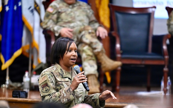 A female military service member in OCP (Operational Camouflage Pattern) uniform is speaking into a microphone at what appears to be a formal military briefing or town hall event. She is positioned in front of a stage area where other uniformed personnel are seated in chairs. An audience of military personnel is visible in the foreground.