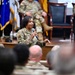 A female military service member in OCP (Operational Camouflage Pattern) uniform is speaking into a microphone at what appears to be a formal military briefing or town hall event. She is positioned in front of a stage area where other uniformed personnel are seated in chairs. An audience of military personnel is visible in the foreground.