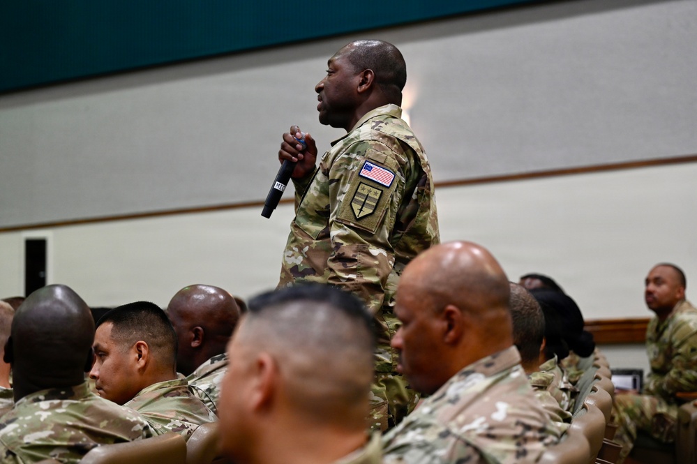 A military service member in OCP (Operational Camouflage Pattern) uniform stands holding a microphone among a seated audience of fellow service members in a large indoor venue. The standing individual appears to be asking a question or making a comment during a briefing, town hall, or similar military gathering. All personnel are wearing the same camouflage uniform pattern with US flag patches visible.