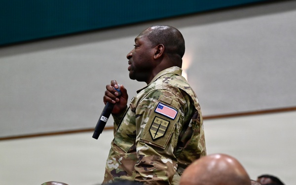 A military service member in OCP (Operational Camouflage Pattern) uniform stands holding a microphone among a seated audience of fellow service members in a large indoor venue. The standing individual appears to be asking a question or making a comment during a briefing, town hall, or similar military gathering. All personnel are wearing the same camouflage uniform pattern with US flag patches visible.