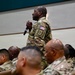 A military service member in OCP (Operational Camouflage Pattern) uniform stands holding a microphone among a seated audience of fellow service members in a large indoor venue. The standing individual appears to be asking a question or making a comment during a briefing, town hall, or similar military gathering. All personnel are wearing the same camouflage uniform pattern with US flag patches visible.