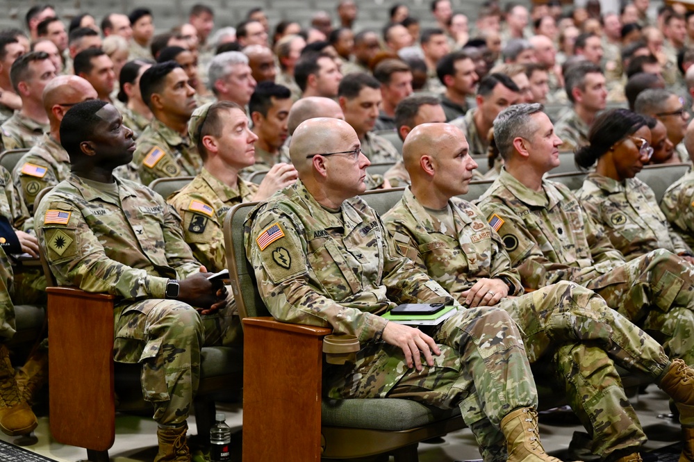 A large group of U.S. Army soldiers in Operational Camouflage Pattern (OCP) uniforms are seated in an auditorium-style venue, attending what appears to be a briefing, ceremony, or training event. The audience fills the visible seating area from front to back, with several senior personnel visible in the foreground. American flag patches and various unit patches are visible on the uniforms.