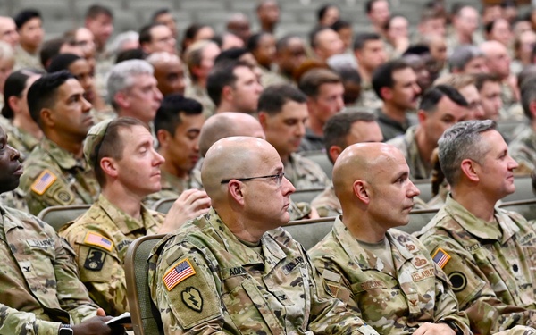 A large group of U.S. Army soldiers in Operational Camouflage Pattern (OCP) uniforms are seated in an auditorium-style venue, attending what appears to be a briefing, ceremony, or training event. The audience fills the visible seating area from front to back, with several senior personnel visible in the foreground. American flag patches and various unit patches are visible on the uniforms.