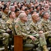 A large group of U.S. Army soldiers in Operational Camouflage Pattern (OCP) uniforms are seated in an auditorium-style venue, attending what appears to be a briefing, ceremony, or training event. The audience fills the visible seating area from front to back, with several senior personnel visible in the foreground. American flag patches and various unit patches are visible on the uniforms.