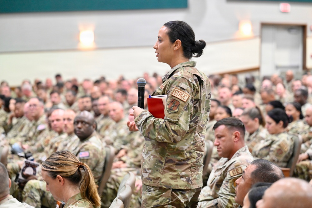 A female military service member stands holding a microphone and a red notebook, appearing to ask a question or make a comment during a large military assembly or briefing. She is surrounded by hundreds of seated uniformed personnel in a large auditorium or meeting hall. The setting suggests a formal all-hands meeting, town hall, or command briefing.