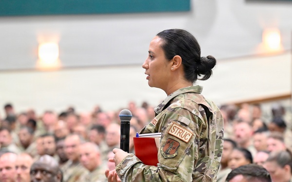 A female military service member stands holding a microphone and a red notebook, appearing to ask a question or make a comment during a large military assembly or briefing. She is surrounded by hundreds of seated uniformed personnel in a large auditorium or meeting hall. The setting suggests a formal all-hands meeting, town hall, or command briefing.