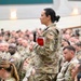 A female military service member stands holding a microphone and a red notebook, appearing to ask a question or make a comment during a large military assembly or briefing. She is surrounded by hundreds of seated uniformed personnel in a large auditorium or meeting hall. The setting suggests a formal all-hands meeting, town hall, or command briefing.