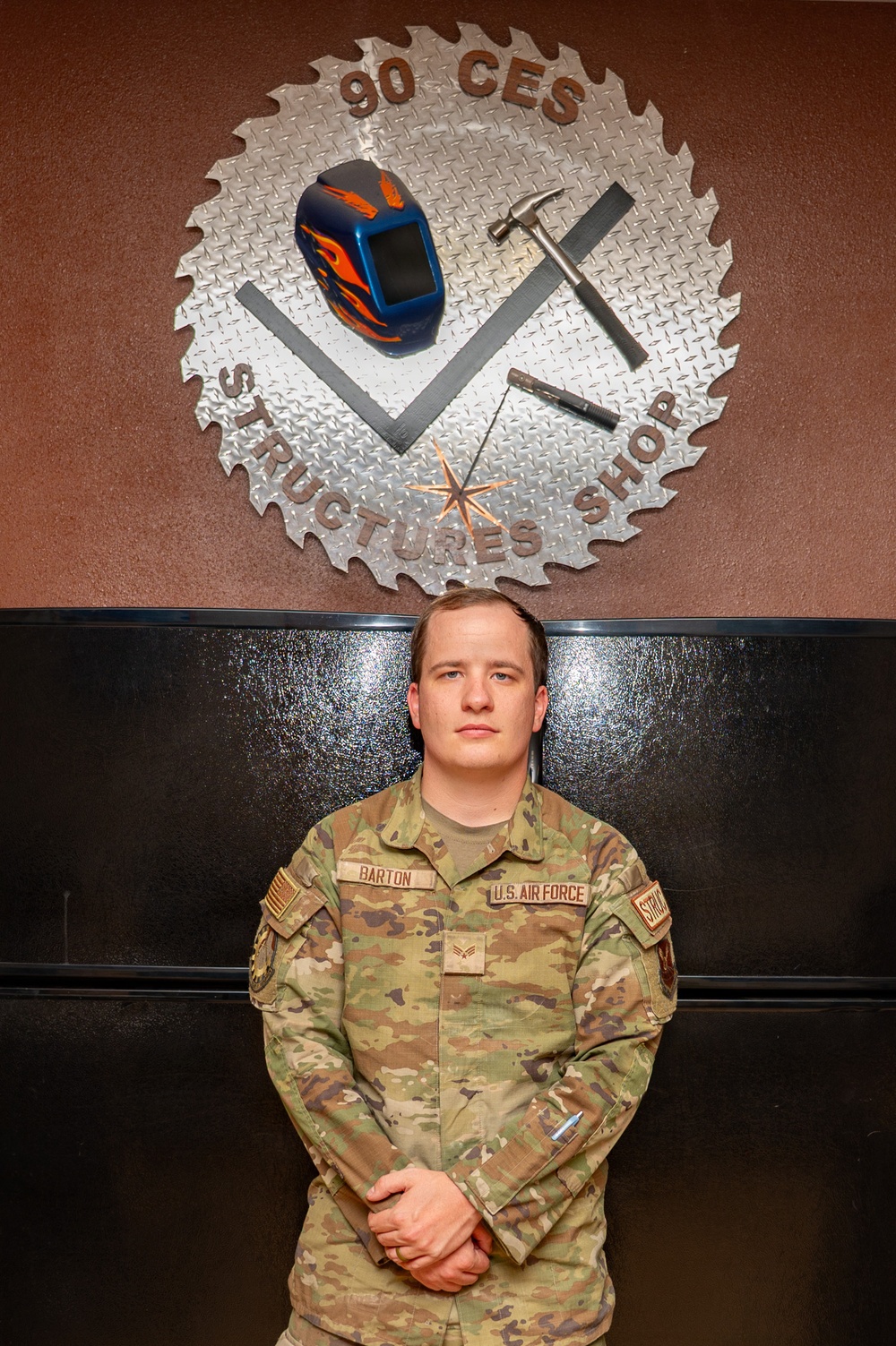 A U.S. Air Force service member in OCP (Operational Camouflage Pattern) uniform stands at parade rest in front of a decorative metal emblem. The name tape reads 'BARTON' and the branch tape reads 'U.S. AIR FORCE.' The emblem behind them identifies the location as the '90 CES Structures Shop,' featuring a saw blade design with tools including a welding helmet, hammer, and chisel.