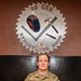 A U.S. Air Force service member in OCP (Operational Camouflage Pattern) uniform stands at parade rest in front of a decorative metal emblem. The name tape reads 'BARTON' and the branch tape reads 'U.S. AIR FORCE.' The emblem behind them identifies the location as the '90 CES Structures Shop,' featuring a saw blade design with tools including a welding helmet, hammer, and chisel.