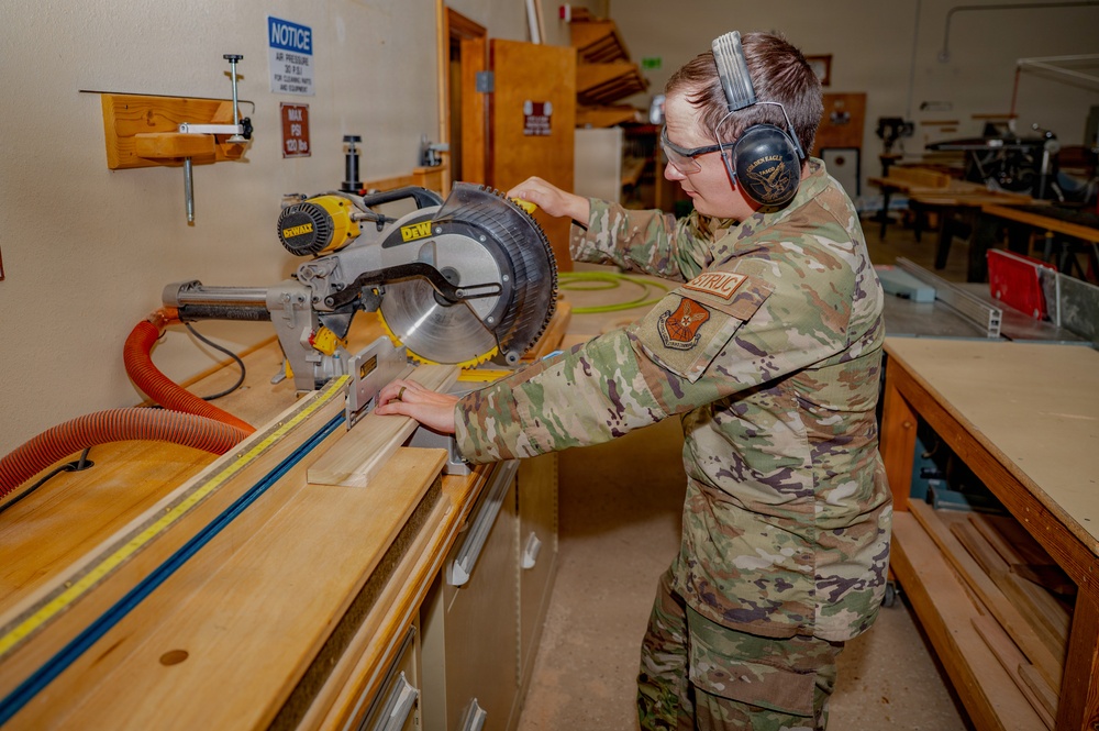 A military service member in OCP (Operational Camouflage Pattern) uniform is operating a DeWalt miter saw in what appears to be a woodworking shop or carpentry facility. The individual is wearing proper personal protective equipment including safety glasses and hearing protection (ear muffs). The setting appears to be a military base workshop or training facility.