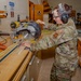 A military service member in OCP (Operational Camouflage Pattern) uniform is operating a DeWalt miter saw in what appears to be a woodworking shop or carpentry facility. The individual is wearing proper personal protective equipment including safety glasses and hearing protection (ear muffs). The setting appears to be a military base workshop or training facility.
