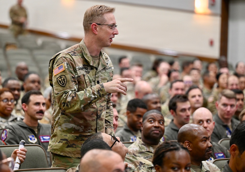 A U.S. Army officer wearing OCP (Operational Camouflage Pattern) uniform is standing and speaking or asking a question during a large military briefing or assembly. The audience consists of a large number of military personnel, predominantly wearing U.S. Air Force flight suits, seated in an auditorium-style venue. The setting appears to be a joint-service gathering or all-hands meeting.