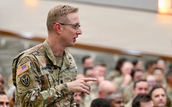 A U.S. Army officer wearing OCP (Operational Camouflage Pattern) uniform is standing and speaking or asking a question during a large military briefing or assembly. The audience consists of a large number of military personnel, predominantly wearing U.S. Air Force flight suits, seated in an auditorium-style venue. The setting appears to be a joint-service gathering or all-hands meeting.