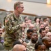 A U.S. Army officer wearing OCP (Operational Camouflage Pattern) uniform is standing and speaking or asking a question during a large military briefing or assembly. The audience consists of a large number of military personnel, predominantly wearing U.S. Air Force flight suits, seated in an auditorium-style venue. The setting appears to be a joint-service gathering or all-hands meeting.