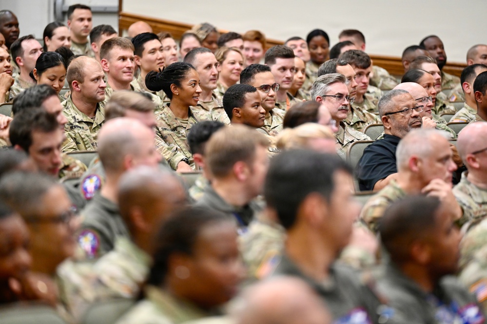 A large group of military personnel, predominantly wearing camouflage uniforms, are seated in an auditorium or briefing hall. The audience appears to be attending a formal military briefing, presentation, or ceremony. The crowd is diverse in age, gender, and ethnicity, with a few individuals in civilian attire visible among the uniformed personnel.