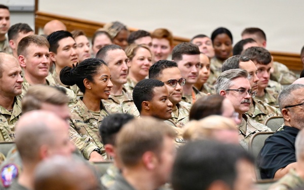 A large group of military personnel, predominantly wearing camouflage uniforms, are seated in an auditorium or briefing hall. The audience appears to be attending a formal military briefing, presentation, or ceremony. The crowd is diverse in age, gender, and ethnicity, with a few individuals in civilian attire visible among the uniformed personnel.
