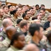 A large group of military personnel, predominantly wearing camouflage uniforms, are seated in an auditorium or briefing hall. The audience appears to be attending a formal military briefing, presentation, or ceremony. The crowd is diverse in age, gender, and ethnicity, with a few individuals in civilian attire visible among the uniformed personnel.