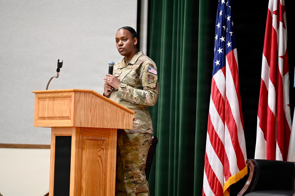 A female military service member in OCP (Operational Camouflage Pattern) uniform is speaking at a wooden podium while holding a microphone. She appears to be delivering a speech or presentation at a formal military event or ceremony. The setting includes an American flag and what appears to be a District of Columbia flag on a stage with green curtain backdrop.