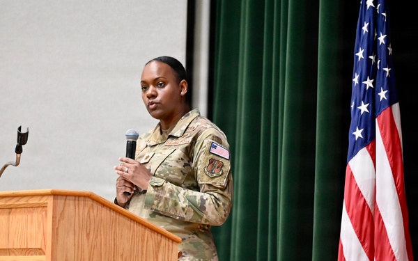 A female military service member in OCP (Operational Camouflage Pattern) uniform is speaking at a wooden podium while holding a microphone. She appears to be delivering a speech or presentation at a formal military event or ceremony. The setting includes an American flag and what appears to be a District of Columbia flag on a stage with green curtain backdrop.