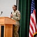 A female military service member in OCP (Operational Camouflage Pattern) uniform is speaking at a wooden podium while holding a microphone. She appears to be delivering a speech or presentation at a formal military event or ceremony. The setting includes an American flag and what appears to be a District of Columbia flag on a stage with green curtain backdrop.