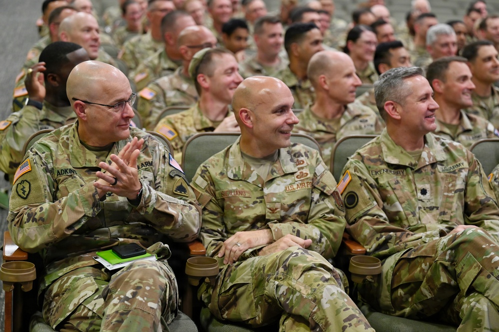 A large group of military personnel in OCP (Operational Camouflage Pattern) uniforms are seated in an auditorium or briefing room, appearing engaged and smiling. The front row includes individuals with visible name tapes reading 'ADKINS,' 'SICILIANO,' and 'EIKENBERRY,' representing both U.S. Army and U.S. Air Force personnel. The atmosphere appears relaxed and positive, suggesting a formal but lighthearted military gathering or ceremony.