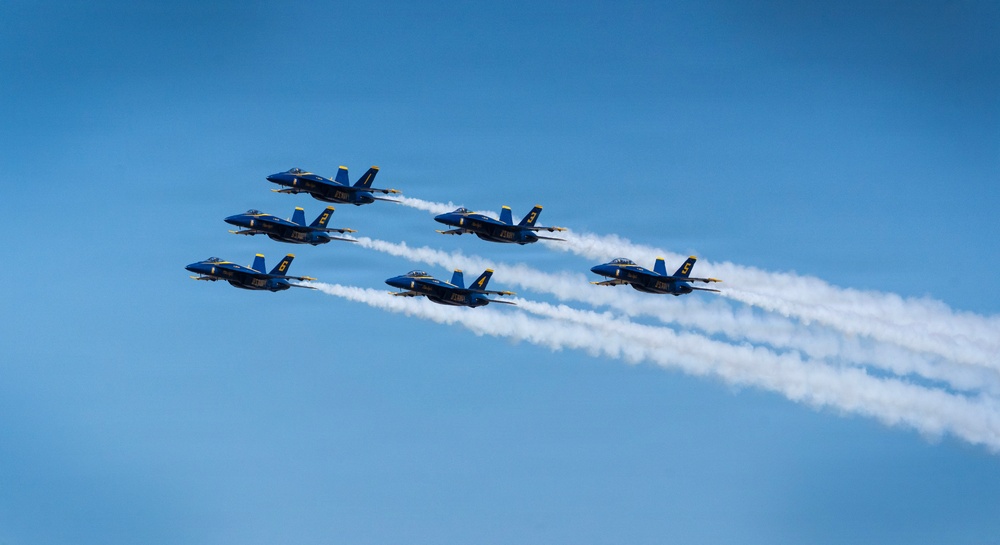 Six U.S. Navy Blue Angels F/A-18 Hornet jets fly in a tight diamond formation against a clear blue sky. The aircraft are producing white smoke trails as part of an aerobatic airshow demonstration. The formation appears to be traveling from left to right in a coordinated, precise maneuver.