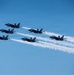 Six U.S. Navy Blue Angels F/A-18 Hornet jets fly in a tight diamond formation against a clear blue sky. The aircraft are producing white smoke trails as part of an aerobatic airshow demonstration. The formation appears to be traveling from left to right in a coordinated, precise maneuver.