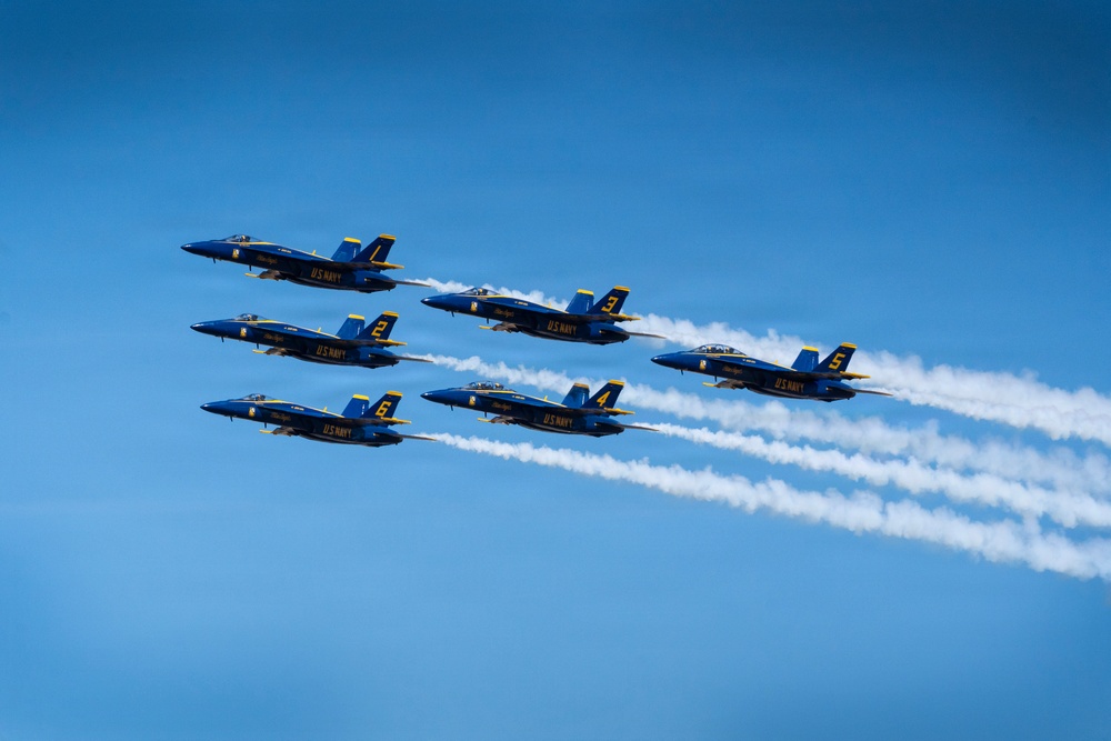 Six U.S. Navy Blue Angels F/A-18 Hornet jets fly in a tight diamond formation against a clear blue sky. The aircraft are numbered 1 through 6 and are emitting white smoke trails as part of an aerial demonstration. The formation is flying in a diagonal orientation from lower-left to upper-right.