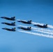 Six U.S. Navy Blue Angels F/A-18 Hornet jets fly in a tight diamond formation against a clear blue sky. The aircraft are numbered 1 through 6 and are emitting white smoke trails as part of an aerial demonstration. The formation is flying in a diagonal orientation from lower-left to upper-right.