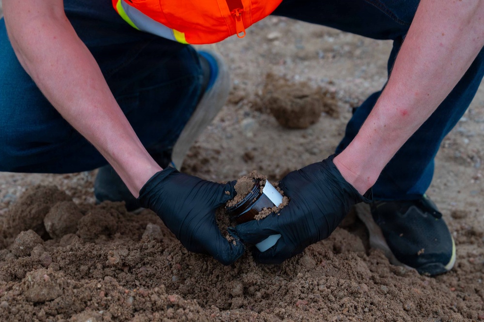 A worker wearing black nitrile gloves and a high-visibility orange safety vest is collecting a soil sample into a small amber/brown sample jar at a construction or environmental testing site. The person is crouching down close to the ground, pressing the sample container into loose, sandy-brown soil. This appears to be an environmental soil sampling or geotechnical testing procedure.