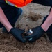 A worker wearing black nitrile gloves and a high-visibility orange safety vest is collecting a soil sample into a small amber/brown sample jar at a construction or environmental testing site. The person is crouching down close to the ground, pressing the sample container into loose, sandy-brown soil. This appears to be an environmental soil sampling or geotechnical testing procedure.