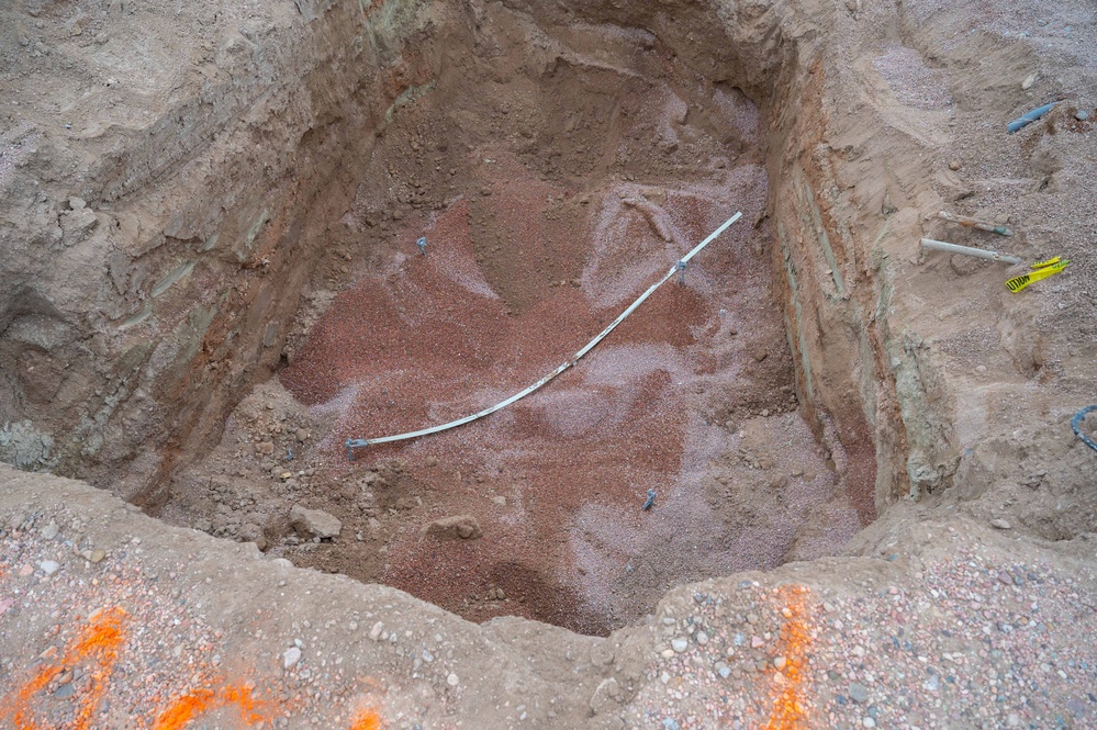 An excavated pit or trench showing exposed soil layers with a curved white conduit or pipe running diagonally across the bottom. The excavation reveals distinct soil strata on the walls, and a reddish-pink granular material (likely sand or gravel bedding) is spread across the pit floor. Survey markers and caution tape are visible along the right wall edge.