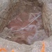 An excavated pit or trench showing exposed soil layers with a curved white conduit or pipe running diagonally across the bottom. The excavation reveals distinct soil strata on the walls, and a reddish-pink granular material (likely sand or gravel bedding) is spread across the pit floor. Survey markers and caution tape are visible along the right wall edge.