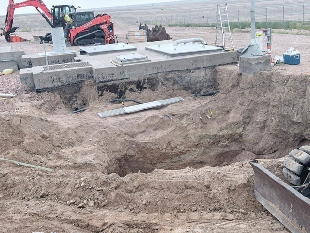 An active construction site showing a large excavation pit with exposed sandy/clay soil. Precast concrete structures, likely utility vaults or junction boxes with metal access hatches, are visible at the edge of the excavation. Heavy equipment and construction materials are staged in the background.