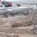 An active construction site showing a large excavation pit with exposed sandy/clay soil. Precast concrete structures, likely utility vaults or junction boxes with metal access hatches, are visible at the edge of the excavation. Heavy equipment and construction materials are staged in the background.