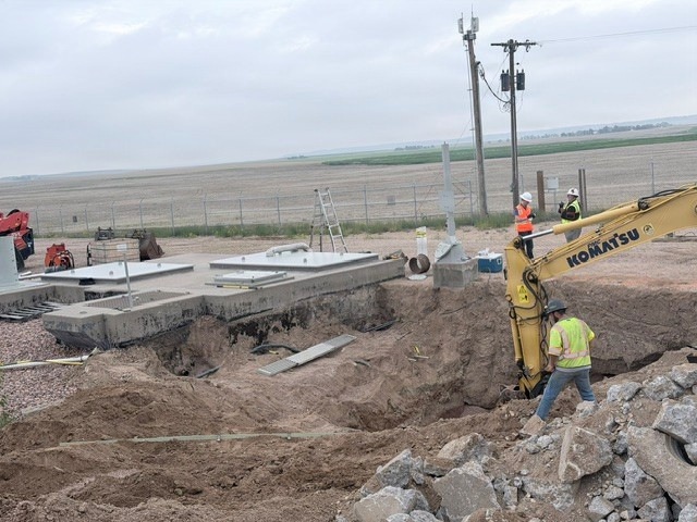 A construction site in a flat, rural landscape showing active excavation work. A yellow Komatsu excavator is operating near a large open trench, with concrete structures and infrastructure already in place. Multiple workers in high-visibility safety gear are present on site.