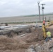 A construction site in a flat, rural landscape showing active excavation work. A yellow Komatsu excavator is operating near a large open trench, with concrete structures and infrastructure already in place. Multiple workers in high-visibility safety gear are present on site.