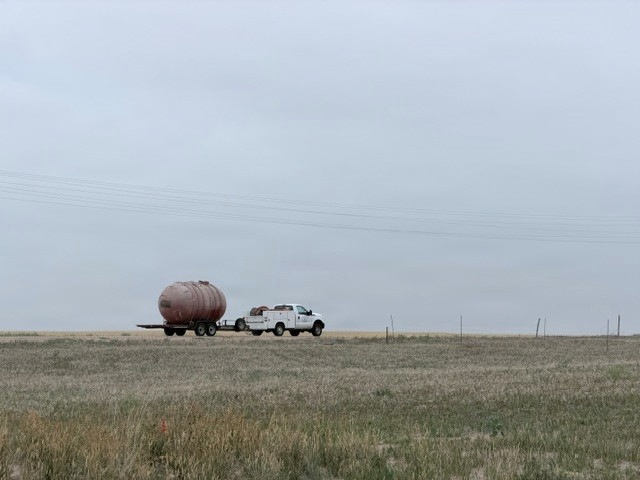 A white pickup truck with a utility bed is towing a large spherical or oval-shaped tank on a flatbed trailer across an open prairie or grassland. The scene appears to be in a rural agricultural or oil/gas region under overcast skies. Power lines run diagonally across the background, and fence posts are visible along the horizon.