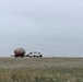 A white pickup truck with a utility bed is towing a large spherical or oval-shaped tank on a flatbed trailer across an open prairie or grassland. The scene appears to be in a rural agricultural or oil/gas region under overcast skies. Power lines run diagonally across the background, and fence posts are visible along the horizon.