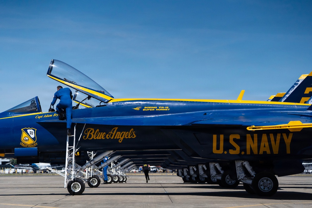 A U.S. Navy Blue Angels Boeing F/A-18 Super Hornet is shown on the tarmac during what appears to be an airshow. The aircraft displays the iconic blue and gold color scheme with 'Blue Angels' and 'U.S. NAVY' markings prominently visible on the fuselage. Ground crew personnel are actively servicing the aircraft, with one individual climbing into the cockpit via a ladder.