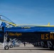 A U.S. Navy Blue Angels Boeing F/A-18 Super Hornet is shown on the tarmac during what appears to be an airshow. The aircraft displays the iconic blue and gold color scheme with 'Blue Angels' and 'U.S. NAVY' markings prominently visible on the fuselage. Ground crew personnel are actively servicing the aircraft, with one individual climbing into the cockpit via a ladder.