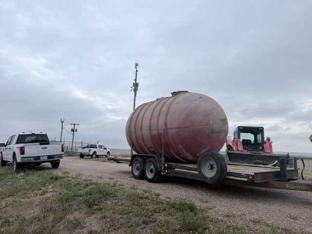 The image shows a rural, open landscape with a large cylindrical storage tank loaded on a dual-axle flatbed trailer, branded 'Kaufman'. Two white Ford pickup trucks are visible in the background near what appears to be a fenced facility. The scene suggests an industrial or agricultural equipment transport operation under overcast skies.