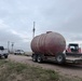 The image shows a rural, open landscape with a large cylindrical storage tank loaded on a dual-axle flatbed trailer, branded 'Kaufman'. Two white Ford pickup trucks are visible in the background near what appears to be a fenced facility. The scene suggests an industrial or agricultural equipment transport operation under overcast skies.