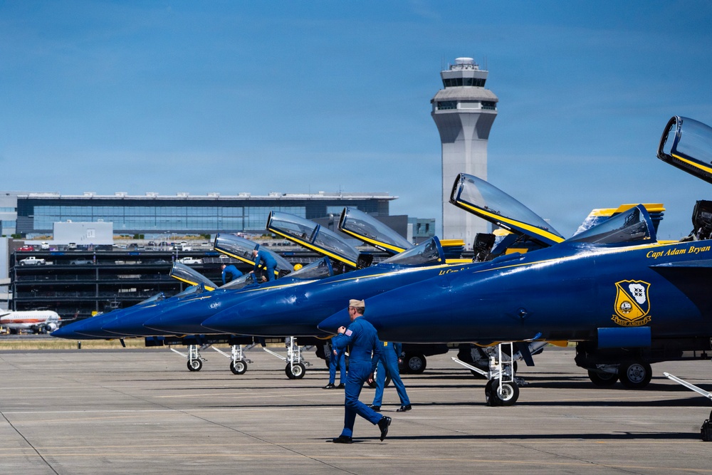 A row of U.S. Navy Blue Angels F/A-18 Hornet jets are parked on an airport tarmac, identifiable by their distinctive blue and gold paint scheme. Personnel in blue flight suits are walking near and climbing on the aircraft. A large airport control tower and terminal building are visible in the background, suggesting this is a major civilian or joint-use airport.
